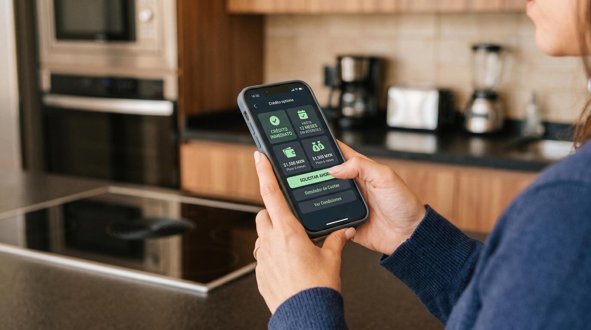 Primer plano de manos sosteniendo un teléfono celular mostrando opciones de crédito en la pantalla, con electrodomésticos de cocina modernos desenfoca